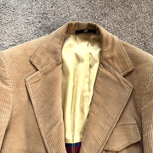 May include: A tan corduroy blazer featuring a notched lapel, a light yellow interior lining, and a small dark tag at the inner collar. The garment displays a textured ribbed fabric surface and a glimpse of a red and blue plaid pattern at the hem.