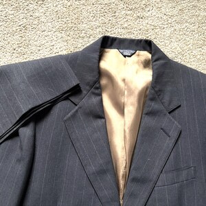 May include: A dark gray pinstripe blazer with a gold-colored interior lining. The blazer has a notched lapel, a single-breasted closure, and a pocket on the left side. The sleeve is folded over, showing the pinstripe pattern.
