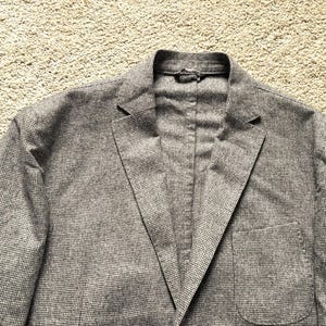 Puede incluir: Una chaqueta gris con un sutil estampado a cuadros. La chaqueta tiene un cierre de botón único y un bolsillo en el lado izquierdo.