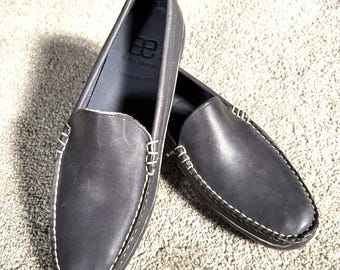 Könnte beinhalten: Ein Paar dunkelgraue Leder-Loafer mit weißen Nähten. Die Schuhe haben eine runde Spitze und ein Slip-on-Design. Die Innenseite des Schuhs ist hellgrau. Der Text "ae by Alan Edmonds Interstate 90" ist sichtbar.