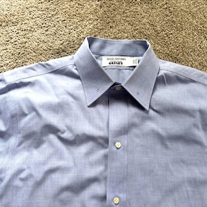 Puede incluir: Una camisa azul claro con cuello clásico y cierre de botones. La camisa tiene las etiquetas "Brooks Brothers" y "GATSBY" en el cuello. La tela parece ser de algodón tejido o una mezcla.