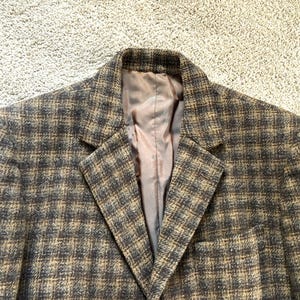 May include: A close-up of a tweed blazer in a brown and gray plaid pattern. The blazer has a notched lapel and a brown satin lining. The fabric has a textured appearance.