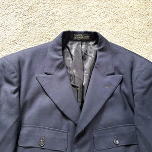 Könnte beinhalten: Ein marineblauer Blazer mit fallendem Revers, zwei Vordertaschen und einem Zwei-Knopf-Verschluss. Das Innenfutter ist schwarz. Der Blazer ist aus einem einfarbigen, dunkelblauen Stoff gefertigt.