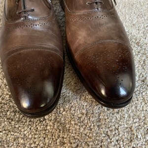 May include: A pair of brown leather Oxford shoes with a brogue design. The shoes have a lace-up closure and a cap toe.