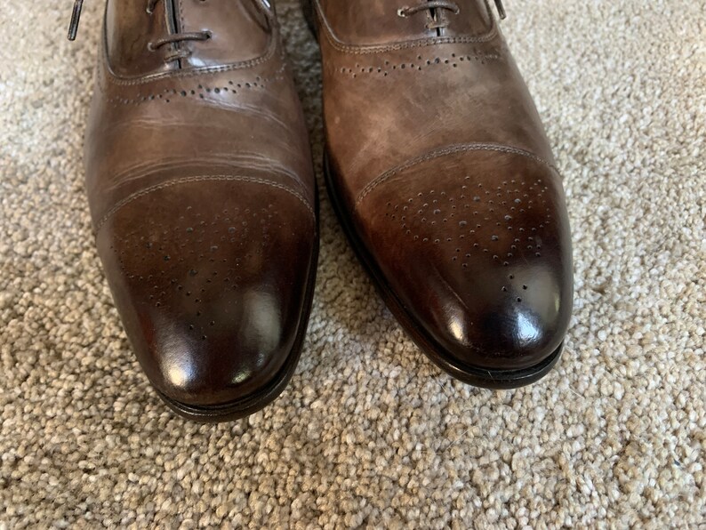 May include: A pair of brown leather Oxford shoes with a brogue design. The shoes have a lace-up closure and a cap toe.