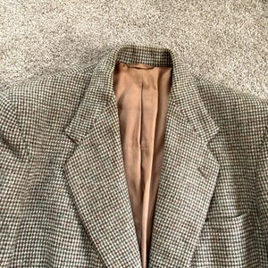 May include: A blazer featuring a brown and tan houndstooth pattern with a textured weave. The garment has notched lapels and a solid tan lining visible at the front opening. The structure includes a single button closure and a chest pocket.