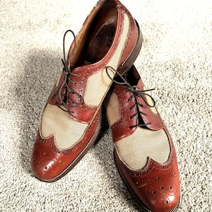 Può includere: Un paio di scarpe stringate wingtip in pelle marrone e beige con un pannello in tessuto intrecciato. Le scarpe hanno lacci marrone scuro e un design classico, adatte per occasioni formali o semi-formali.