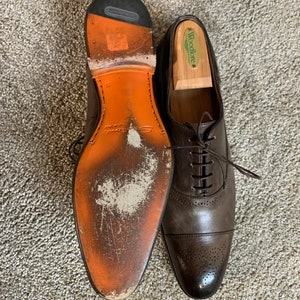 May include: A pair of brown leather Oxford shoes with a lace-up fastening. The shoes feature a cap toe design and a perforated pattern on the toe. One shoe has a wooden shoe tree inside and the other shoe has a worn orange leather sole.