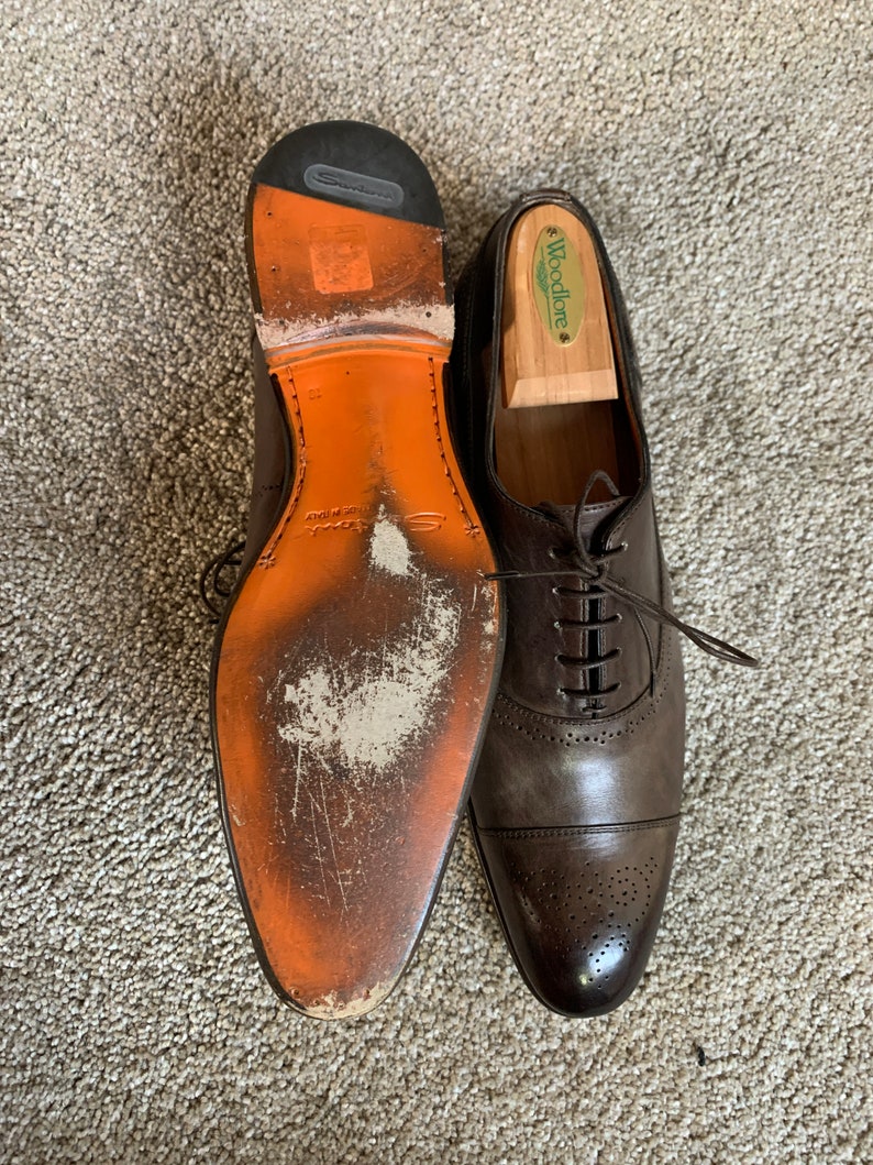 May include: A pair of brown leather Oxford shoes with a lace-up fastening. The shoes feature a cap toe design and a perforated pattern on the toe. One shoe has a wooden shoe tree inside and the other shoe has a worn orange leather sole.