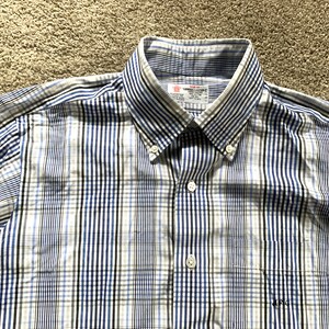 Könnte beinhalten: Ein langärmliges Hemd mit Knöpfen in weiß, blau und schwarzem Karomuster mit einer kleinen Tasche auf der linken Seite. Das Hemd hat ein Etikett mit der Aufschrift "Thomas & Mercer" in Rot.