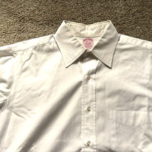 Puede incluir: Primer plano de una camisa blanca abotonada. La camisa tiene cuello puntiagudo, bolsillo en el pecho y botones en la parte delantera. La tela parece lisa, posiblemente de algodón. La etiqueta Brooks Brothers es visible.