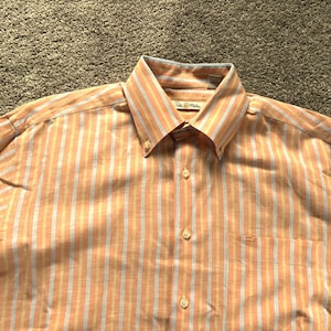 以下が含まれることがあります： オレンジと白のストライプのボタンダウンシャツで、ラベルには「The  Island  Man」と書いてあります。