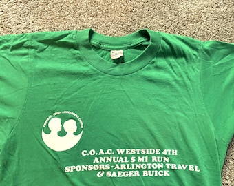 Könnte beinhalten: Grünes T-Shirt mit weißem Text, der "C.O.A.C. Westside 4th Annual 5 Mi Run Sponsors Arlington Travel & Saegar Buick" lautet. Das Shirt hat auch ein weißes Logo mit zwei Kinderköpfen.