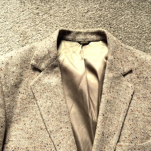 Puede incluir: Un blazer de tweed en una paleta de colores neutros, con motas marrones y beige. La chaqueta tiene solapas con muescas, un diseño de un solo pecho y un forro de satén de color claro. La estética general es clásica y versátil.