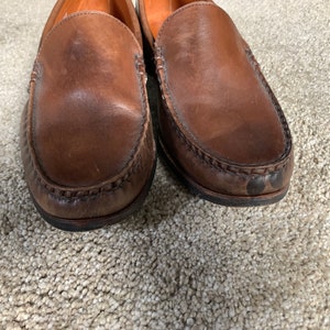 May include: A pair of brown leather loafers with a stitched edge. The loafers have a worn look and are on a light brown carpet.