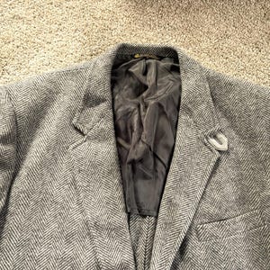 Puede incluir: Un blazer gris con estampado de espiga, solapa con muescas y cierre de un solo botón. El blazer tiene un bolsillo en el pecho y un forro gris. Un pequeño alfiler decorativo está en la solapa.