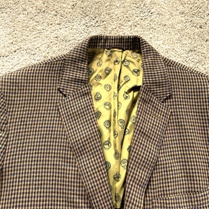 Può includere: Un blazer a quadri marroni e blu con una fodera a motivi gialli. Il blazer ha una chiusura con un bottone e una tasca sul lato sinistro.