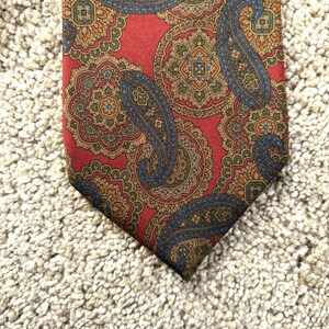 May include: A red tie featuring a paisley pattern in blue, brown, and orange. The intricate design is set against a red background. The tie is made of a smooth material and is displayed on a textured surface.