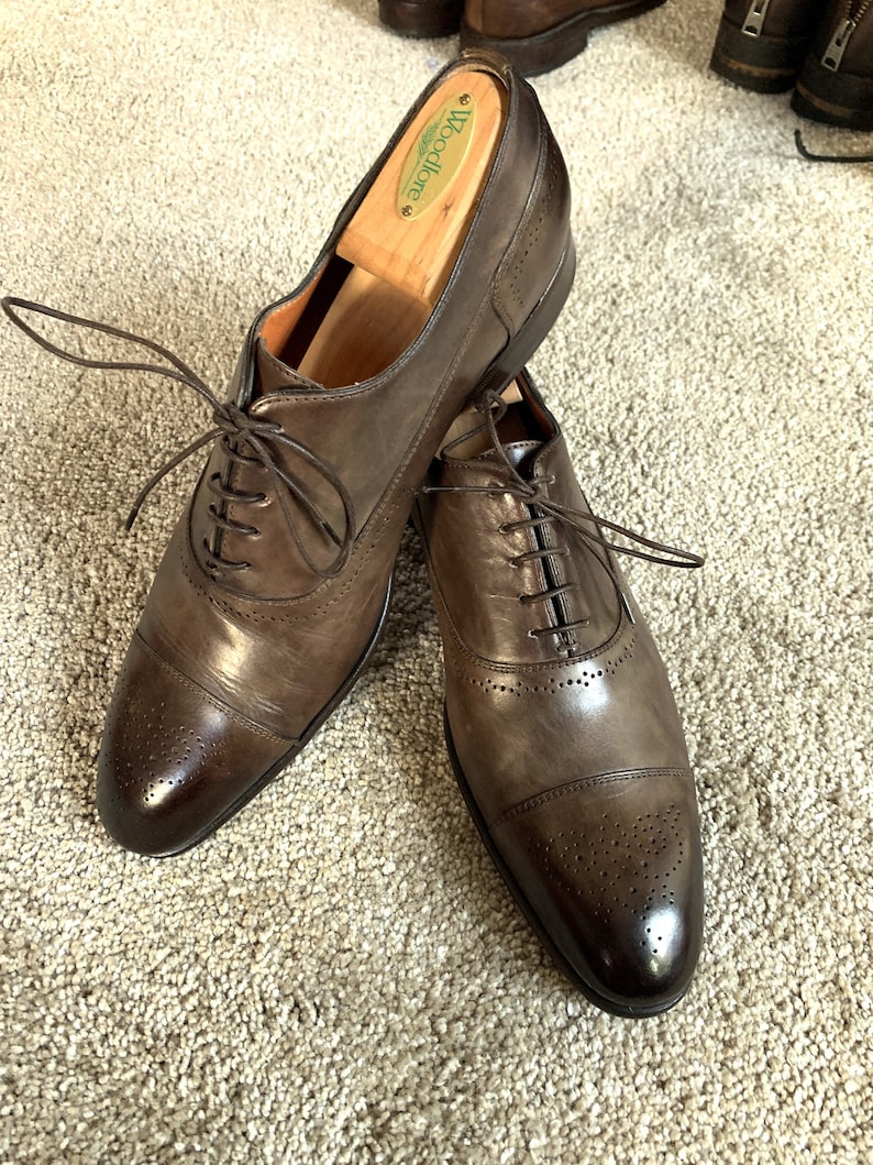 May include: A pair of brown leather Oxford shoes with a cap toe and brogue detailing. The shoes are laced up and have a polished finish. The shoes are on a beige carpet.
