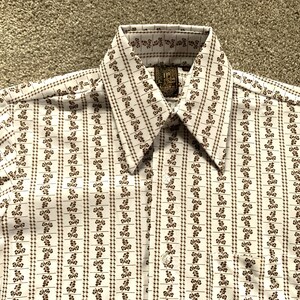 May include: A white button-down shirt with a brown floral pattern. The shirt has a pointed collar and a single button closure. The label on the shirt reads "Goya 20".
