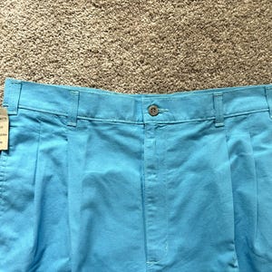 May include: Close-up of light blue pleated shorts with a button closure and belt loops. A tag is attached to the shorts. The shorts are size 32.