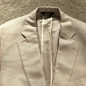 Könnte beinhalten: Ein beiger Blazer mit einem weißen und braunen Hahnentrittmuster. Der Blazer hat einen einzigen Knopfverschluss und ein Etikett mit der Aufschrift "Barneys New York".