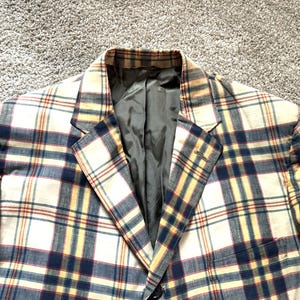 May include: A plaid blazer in cream, navy, and yellow. The jacket has a classic notched lapel and a dark green lining. The pattern is a traditional plaid design, with intersecting lines creating a grid-like pattern.