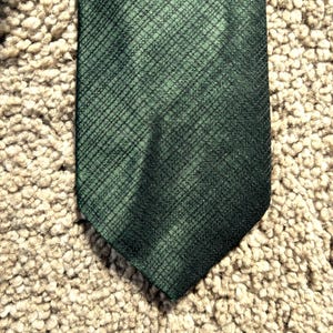 Puede incluir: Una corbata verde oscuro con un sutil patrón de trama cruzada. La corbata está hecha de una tela texturizada y tiene un extremo puntiagudo clásico. El fondo es una superficie texturizada neutra.