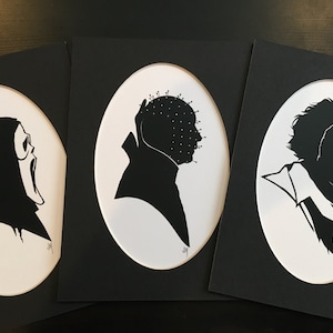 Könnte beinhalten: Drei gerahmte Schwarz-Weiß-Silhouette-Kunstdrucke. Die Drucke zeigen ikonische Horrorfilmfiguren: Ghostface, Pinhead und Beetlejuice. Jede Silhouette ist vor einem weißen ovalen Hintergrund auf einem schwarzen rechteckigen Rahmen angebracht.