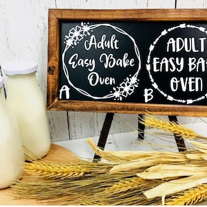 May include: A wooden sign with a chalkboard insert that reads "Adult Easy Bake Oven" in a white circle with floral accents. The sign is on a small easel and there are three glass bottles with white lids in front of the sign.