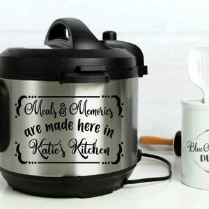 May include: A stainless steel electric pressure cooker with a black lid and a black decal that reads "{ Meals & Memories are made here in Katie's Kitchen }"