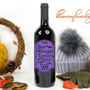 May include: A purple and black wine bottle label with the text "Bat Wings Tonic Elixir Recipe #666" and a bat graphic. The bottle is surrounded by fall decor, including a pumpkin, a wreath, and cinnamon sticks.