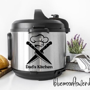 May include: A stainless steel and black electric pressure cooker with a chef hat and crossed knives graphic that reads "Dad's Kitchen". A loaf of bread sits on a wooden cutting board to the left. A small pot of lavender and a striped towel are to the right.