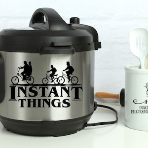 May include: A stainless steel Instant Pot with a black lid and a black decal that says "Instant Things" with a graphic of three people riding bicycles.