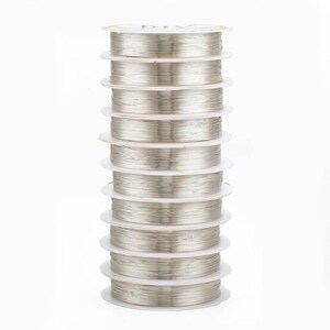 May include: A stack of ten spools of silver-colored beading wire. Each spool is neatly wound and displayed on a white plastic base. The wire appears to be fine and flexible, suitable for jewelry making and other craft projects.