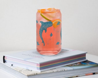 May include: A glass can with a pink drink and fruit inside. The can has a blue and white pattern of manta rays and sharks. The can is sitting on top of two books.