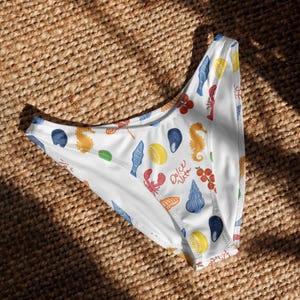 May include: White bikini bottom with a colorful pattern of seafood, lemons, and seahorses. The design includes red lobsters, blue mussels, and the text "Dolce Vita". The swimwear is set against a woven background.