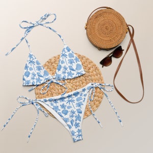 May include: A blue and white floral print bikini with a matching straw bag and sunglasses. The bikini top is a triangle style and the bottom is a tie-side style.