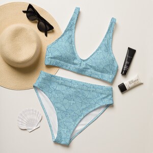 May include: A light blue bikini set with a repeating floral pattern, a wide-brimmed straw hat, and black sunglasses. The set includes a top with a scoop neckline and high-waisted bottoms. Also included are two tubes of sunscreen and a seashell.