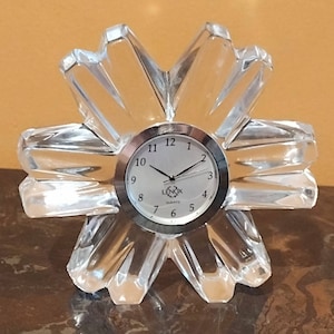 May include: A clear crystal clock with a silver face and black hands. The clock is shaped like a starburst and has the word "LENOX" on the face.
