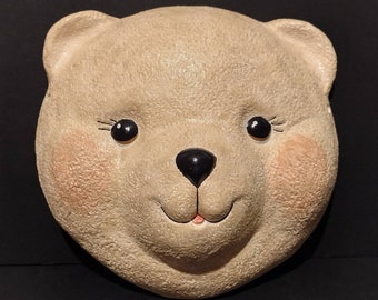 May include: A beige ceramic bear face wall decor with black eyes and a black nose. The bear has a pink tongue sticking out and a slight smile.
