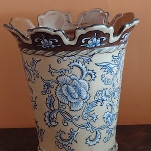 May include: A white ceramic vase with a brown rim and blue floral pattern. The vase has a scalloped top and a rope-like design around the base.