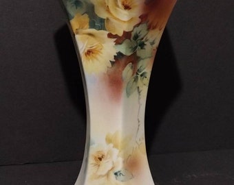 May include: A white ceramic vase with a hexagonal shape and a gold rim. The vase is decorated with a floral pattern of yellow roses and green leaves.