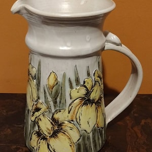 May include: A white ceramic pitcher with a handle and a floral design. The pitcher features yellow daffodils with green leaves and stems.