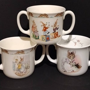 May include: Three white ceramic children's cups with double handles. The cups feature illustrations of bunnies, a mouse, and a bunny playing with blocks that spell ABC.