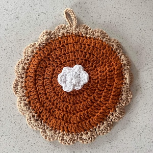 May include: A handmade crochet trivet in the shape of a pie. The trivet features a round design with a brown crust, an orange filling, and a white, cloud-shaped accent in the center. A loop is attached for hanging.