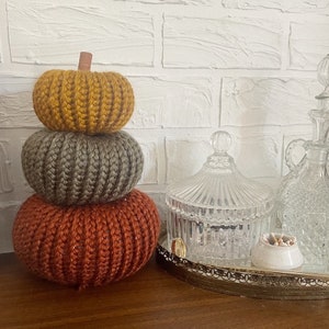 May include: A stack of three knitted pumpkins in orange, gray, and yellow. The pumpkins are stacked on a wooden surface in front of a white brick wall.
