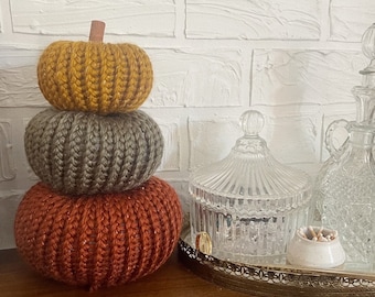 May include: A stack of three knitted pumpkins in orange, gray, and yellow. The pumpkins are stacked on a wooden surface in front of a white brick wall.