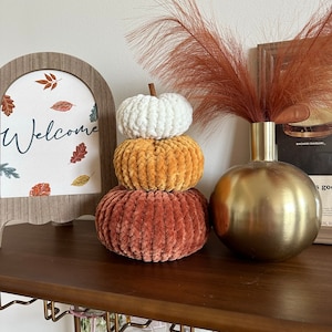 May include: A set of three knitted pumpkins in shades of white, orange, and rust. The pumpkins are stacked on top of each other and are sitting on a wooden shelf. There is a sign that says "Welcome" and a vase with dried pampas grass in the background.