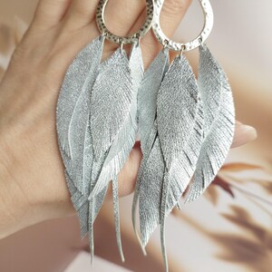 May include: Silver feather earrings with hammered metal teardrop-shaped tops. The earrings feature multiple silver-colored feather-shaped pendants. The earrings are held up by a hand.